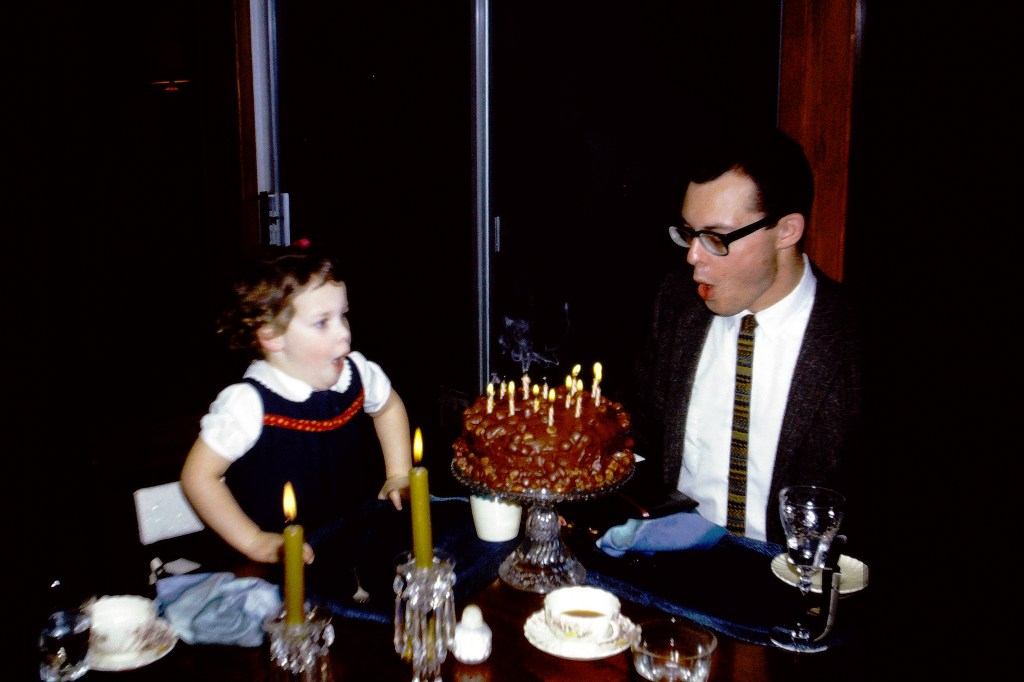 Young man in suit and tie blowing birthday candles across from toddler in blue dress.
