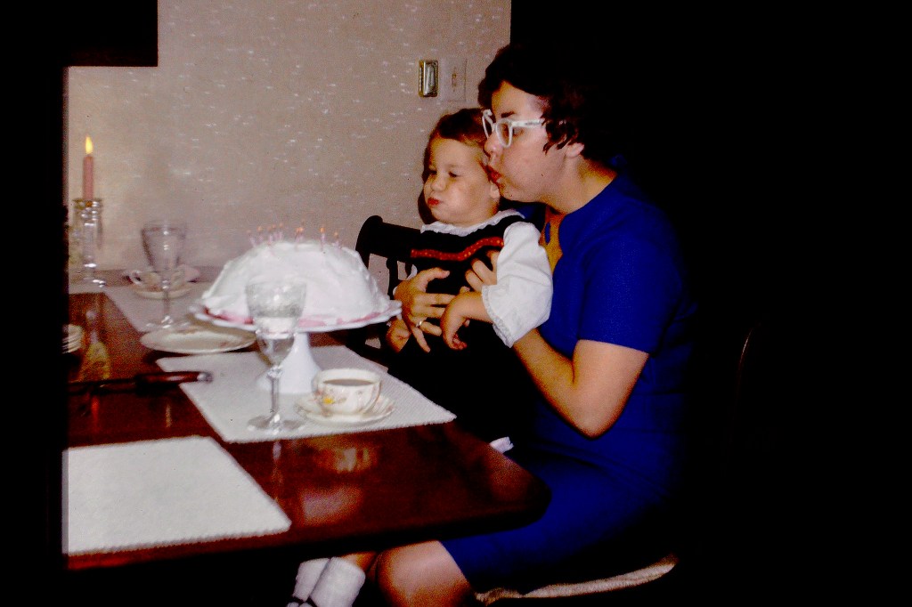 Young woman in blue dress blowing birthday candles and holding toddler.